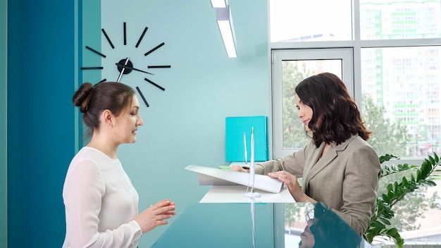 Administrador de mujer positiva en blanco habla con el cliente de pie cerca del mostrador del espejo en la recepción en la vista lateral cercana del hospital contemporáneo