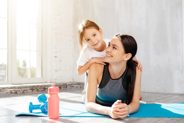 Admirável fêmea descansando no tapete de fitness com seu filho nas costas depois do treino em casa