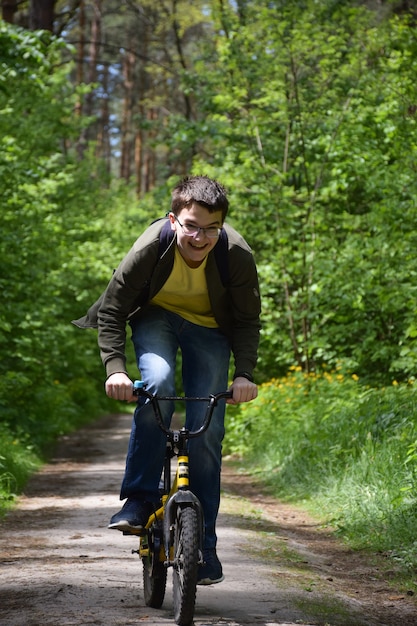 Un adolescente de unos 14 años que usa anteojos anda rápidamente en una bicicleta para niños por un sendero del bosque y se ríe. Vista frontal