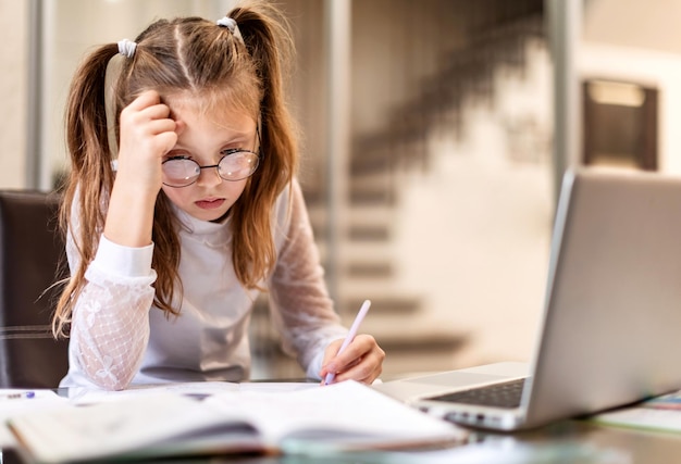 Una adolescente aburrida, cansada de aprender en línea y estudiar haciendo deberes usando una laptop en casa. Educación en el hogar y educación a distancia