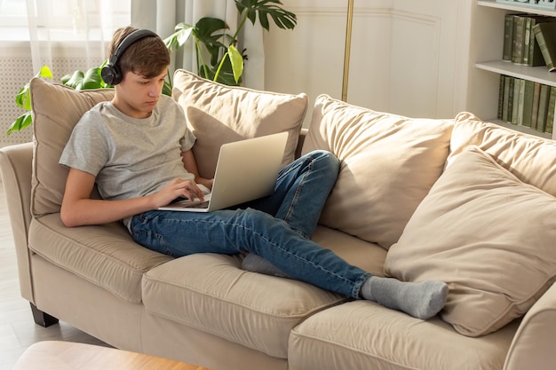 Un adolescente acostado en un sofá en una habitación con auriculares negros en la cabeza mira hacia un portátil gris o