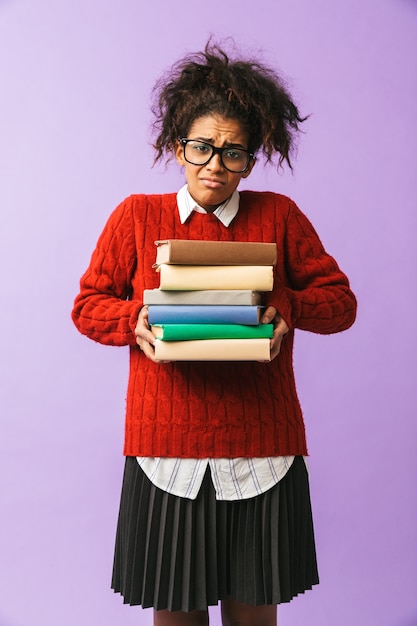 Adolescente afroamericana en uniforme escolar sosteniendo un montón de libros, aislado