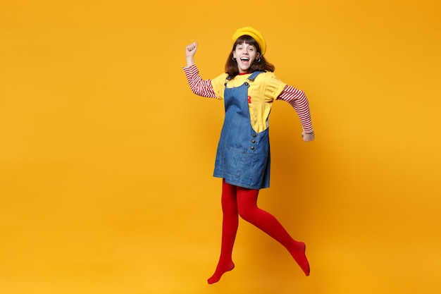 Adolescente alegre con boina francesa, vestido de mezclilla esparciendo las manos, saltando y jugando aislado en el fondo de la pared amarilla en el estudio. Emociones de la gente, concepto de estilo de vida. Simulacros de espacio de copia.