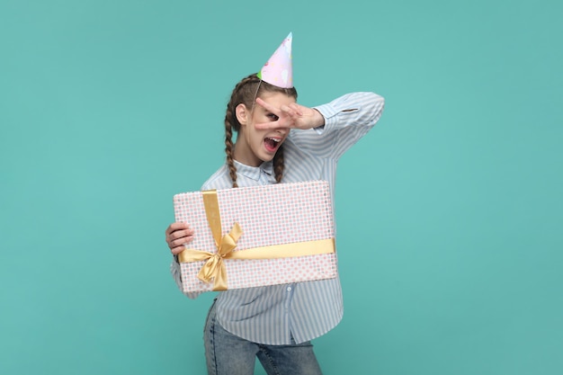 Una adolescente alegre en un cono de fiesta sosteniendo una caja de regalo que muestra el signo de v celebrando su cumpleaños
