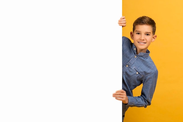 Foto un adolescente alegre espiando desde detrás de un tablero de anuncios blanco en blanco