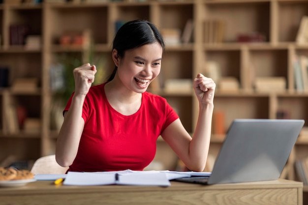 Una adolescente alegre, una estudiante coreana, ve una lección en línea en una computadora portátil y se regocija en la victoria y celebra una gran noticia