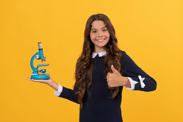 Una adolescente alegre sostiene un microscopio para la educación sobre un fondo amarillo que muestra la química con el pulgar hacia arriba