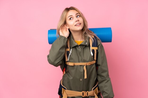 Adolescente alpinista ruso chica con una gran mochila aislada en rosa escuchando algo poniendo la mano en la oreja