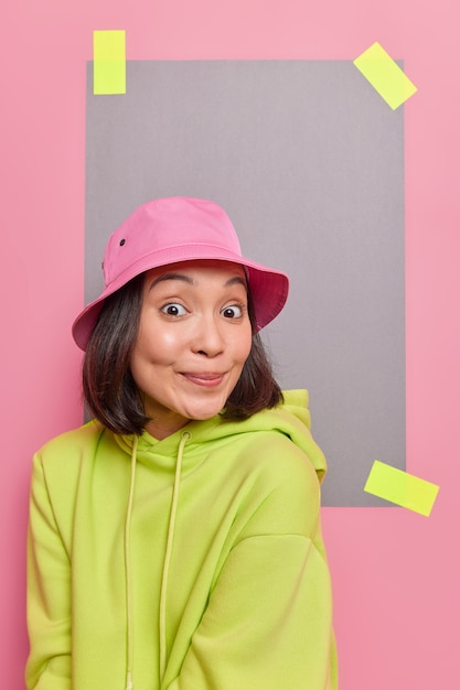 Adolescente con apariencia oriental sonríe alegremente escucha algo con curiosa expresión alegre viste una sudadera con capucha casual aislada sobre una pared rosa