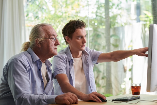 Adolescente apontando para a tela do laptop ao explicar ao avô como usar o aplicativo para comunicação on-line