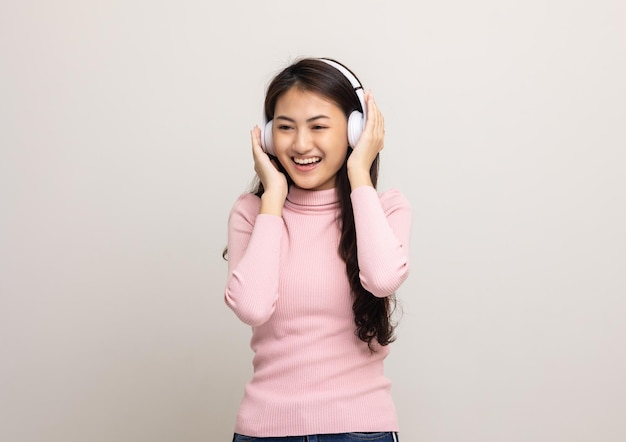 Una adolescente asiática alegre escucha la música con auriculares blancos aislados Hermosa mujer joven con camisa rosa toca un auricular inalámbrico divirtiéndose con la música en la habitación