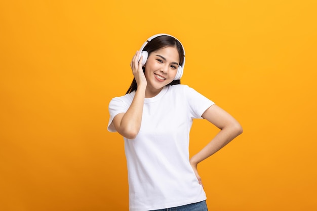 Una adolescente asiática alegre escucha la música con auriculares blancos bailando sobre un fondo amarillo aislado. Hermosa joven en la mano toca un auricular inalámbrico divirtiéndose con la música.