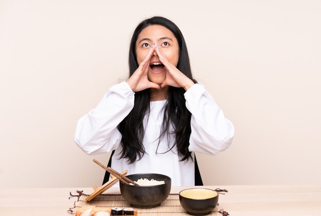 Adolescente asiática comiendo comida asiática en pared beige gritando y anunciando algo