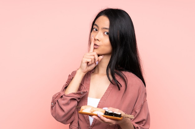 Adolescente asiática comiendo sushi aislado en rosa haciendo gesto de silencio