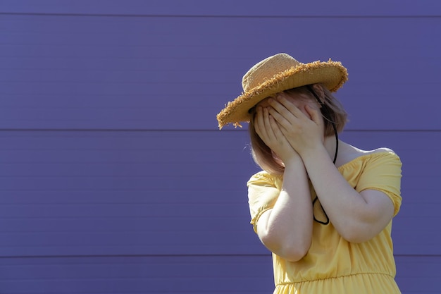 Una adolescente asiática frustrada con un sombrero y un vestido se cubre la cara con las manos en un fondo morado