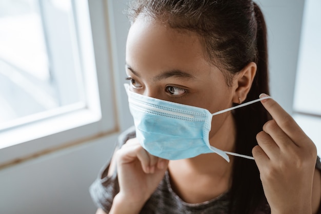 Adolescente asiática lleva una máscara médica mientras está sentada junto a la ventana