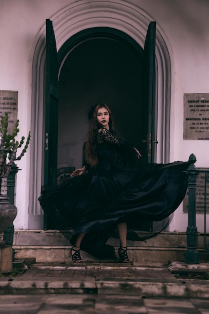 Una adolescente asiática posando con su vestido negro flotando en el aire mientras está de pie frente a un cementerio con todo el maquillaje negro en la cara