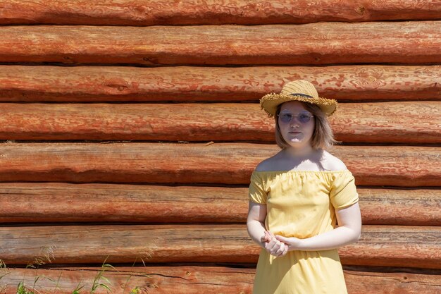 Adolescente asiática con un sombrero de vestir amarillo y gafas de sol cerca de la pared de una casa de pueblo de madera