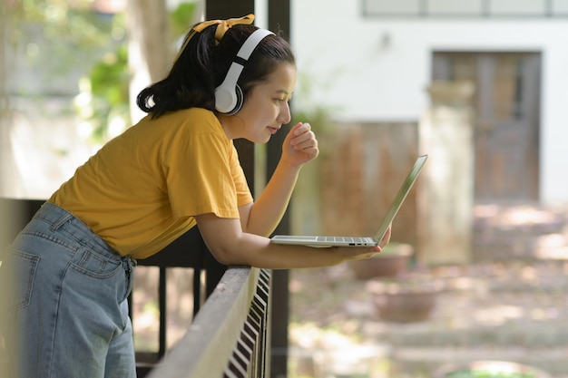 Adolescente asiática usando laptop para trabalhar em casa, garota usando fones de ouvido e em pé usando laptop na varanda, vista lateral, trabalho em casa, escritório em casa, trabalho remoto.