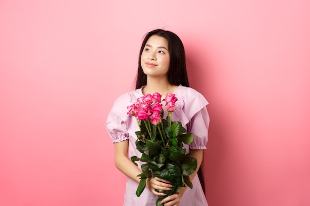 Adolescente asiática en vestido lindo que parece romántico en el logotipo del espacio vacío, con regalo de flores del día de San Valentín, recibe boquet de rosas del amante, de pie sobre fondo rosa.