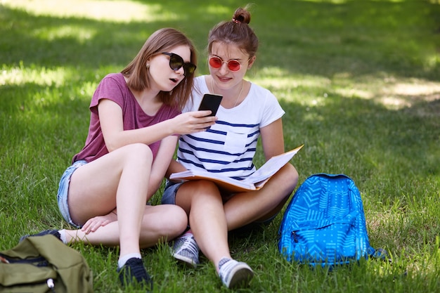 Una adolescente de aspecto agradable sostiene un teléfono inteligente, mira la pantalla junto con un amigo, hace compras en línea mientras descansa en la hierba verde