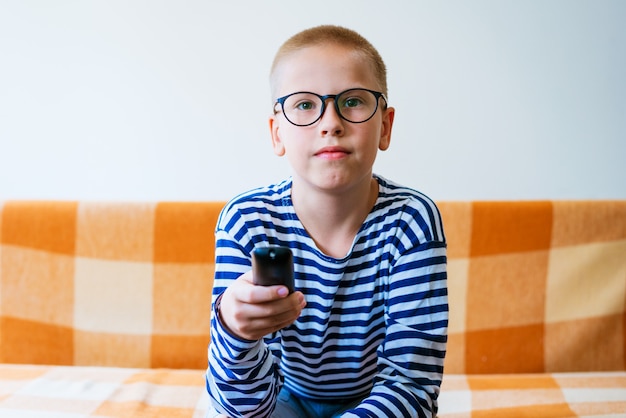 Adolescente assistindo tv com óculos usando o controle remoto enquanto está sentado no sofá na sala de estar em casa em