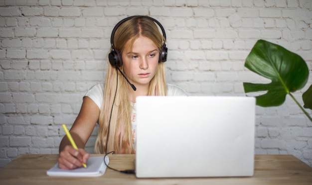 Adolescente en auriculares estudiando en línea y tomando notas en el concepto de notebook, e-learning y educación en el hogar