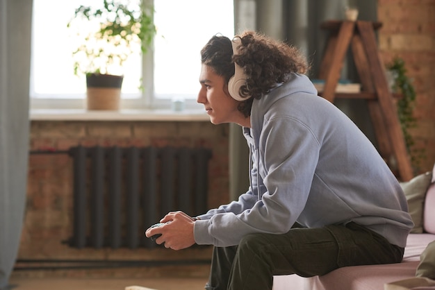 Foto adolescente en auriculares inalámbricos sentado en el sofá y jugando videojuegos en la habitación