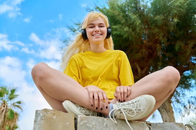 Adolescente en auriculares sentado al aire libre en el cielo azul del parque con nubes