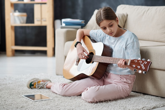 Adolescente autodidacta sentada en una alfombra y usando videos instructivos en tableta mientras toca la guitarra