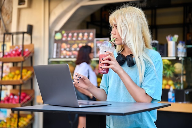 Adolescente en la barra de jugo de fruta al aire libre de frutas bebiendo jugo fresco de vitamina usando una computadora portátil