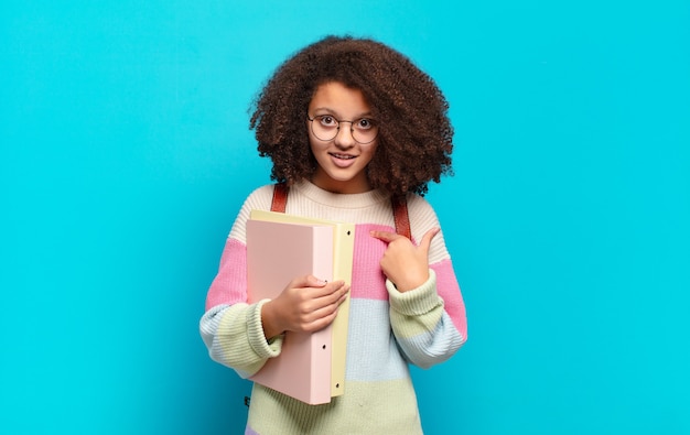 Adolescente bastante afro se sentindo feliz, surpreso e orgulhoso, apontando para si mesmo com um olhar animado e surpreso. conceito de estudante