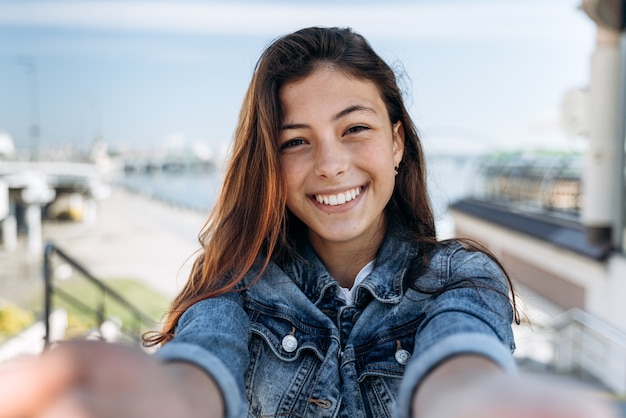 Adolescente bonito e positivo segurando uma câmera, olhando diretamente. Linda garota sorrindo sinceramente no fundo da cidade