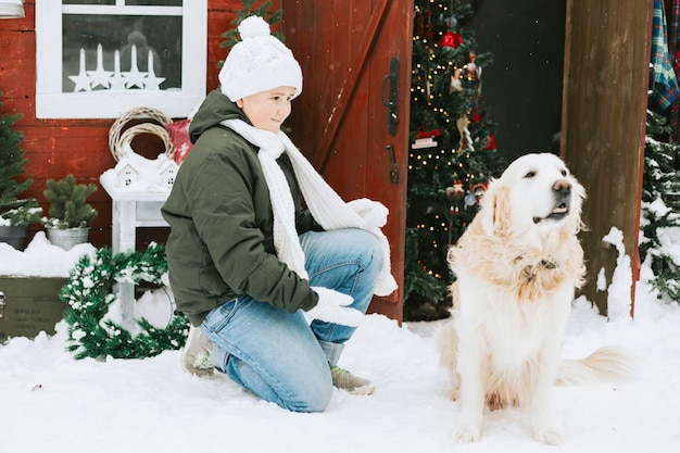 Adolescente bonito em suéter de malha e chapéu se divertindo com a primeira neve e cachorro de estimação fofo labrador