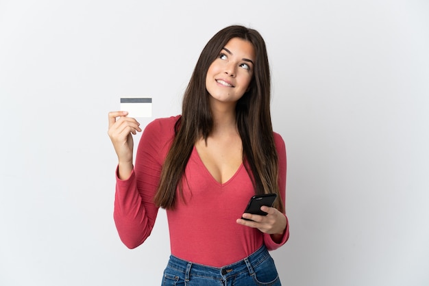 Foto adolescente brasileña aislada en blanco comprando con el móvil con tarjeta de crédito mientras piensa