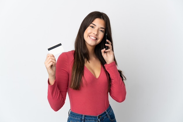 Adolescente brasileña aislada en la pared blanca manteniendo una conversación con el teléfono móvil y sosteniendo una tarjeta de crédito