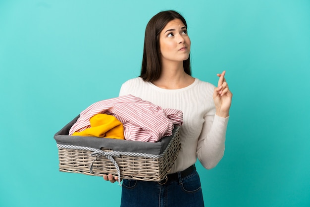 Adolescente brasileña sosteniendo una canasta de ropa aislado sobre fondo azul con los dedos cruzando y deseando lo mejor