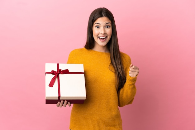 Adolescente brasileña sosteniendo un regalo sobre fondo rosa aislado celebrando una victoria en la posición ganadora