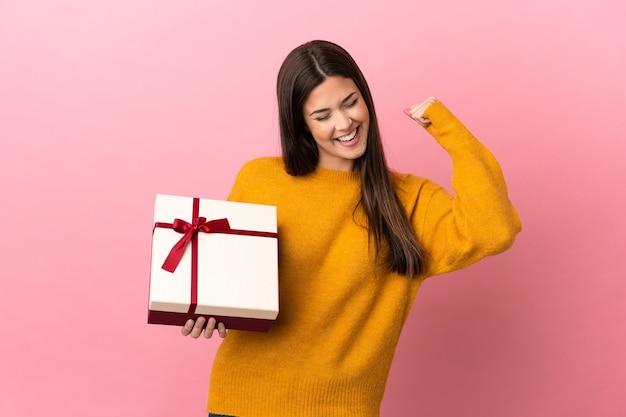 Adolescente brasileña sosteniendo un regalo sobre fondo rosa aislado celebrando una victoria