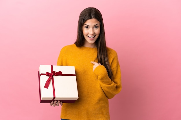 Adolescente brasileña sosteniendo un regalo sobre fondo rosa aislado con expresión facial sorpresa