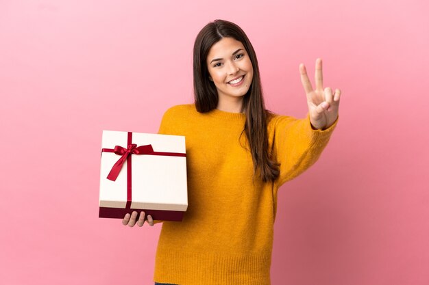 Adolescente brasileña sosteniendo un regalo sobre fondo rosa aislado sonriendo y mostrando el signo de la victoria