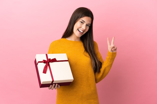 Adolescente brasileña sosteniendo un regalo sobre fondo rosa aislado sonriendo y mostrando el signo de la victoria