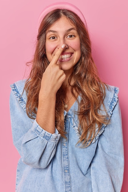 Adolescente brincalhão toca o nariz tem expressão alegre vestida com camisa jeans tenta divertir poses de criança contra estúdio rosa goza tempo livre provoca alguém. Conceito de emoções felizes