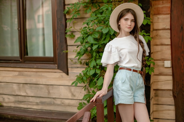 Adolescente de cabello castaño de ensueño con ropa de estilo rural y sombrero de mimbre de pie en el umbral de madera de la casa rural en el día de verano