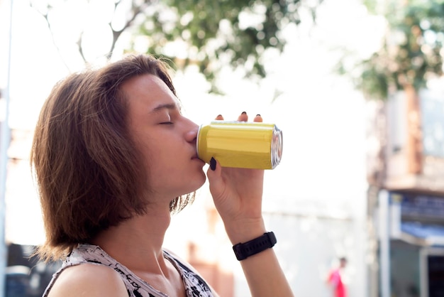 Foto un adolescente con el cabello largo bebe agua gaseosa de una lata una bebida energética en un día caluroso en la calle