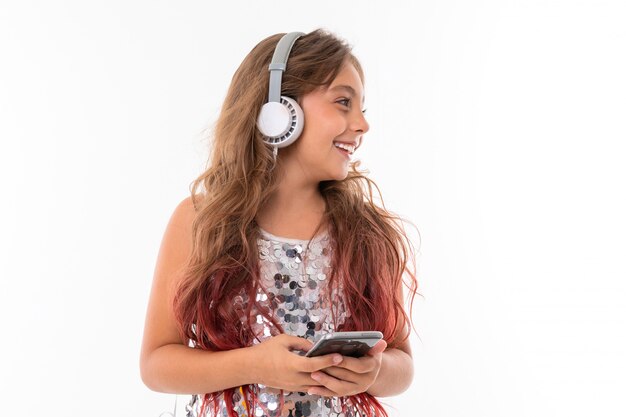 Adolescente con cabello largo y rubio teñido con puntas rosadas, con un vestido claro brillante, de pie con auriculares y sosteniendo el teléfono en la mano