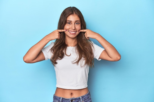 Una adolescente con una camiseta blanca sobre un fondo azul sonríe señalando los dedos a la boca