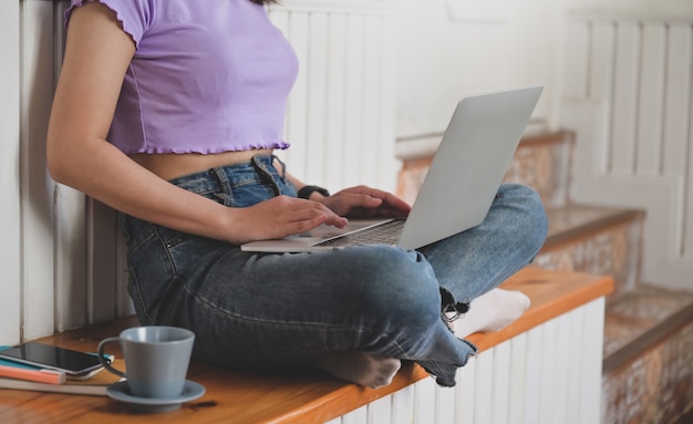 Foto adolescente con una camiseta que muestra su cintura usando una computadora portátil para estudiar en línea desde casa, una mujer que trabaja en una computadora portátil desde casa con una taza de café y un teléfono inteligente.