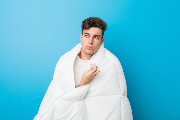 Adolescente cansado homem caucasiano, cobrindo-se com um cobertor e medir sua temperatura corporal