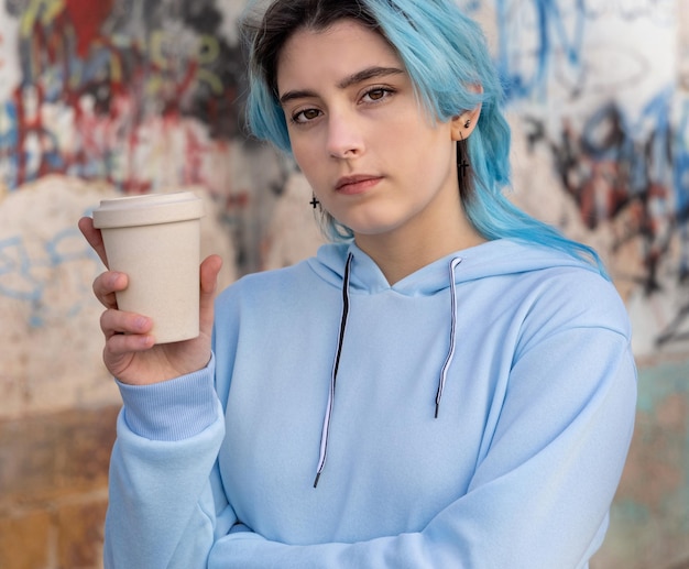 Adolescente con capucha azul claro de gran tamaño con café para llevar y mirando a un lado Adolescente de cabello azul que se queda al aire libre cerca de la pared de graffiti Maqueta de ropa Hipster y concepto de adolescencia